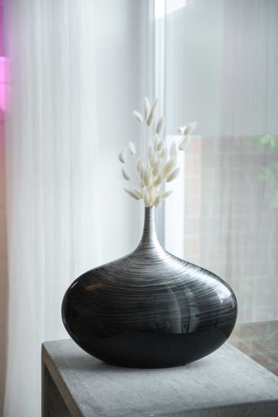 Moderne, bauchige Vase mit feiner Streifenstruktur und getrockneten Gräsern auf einem Steinsockel in einem hellen Raum.