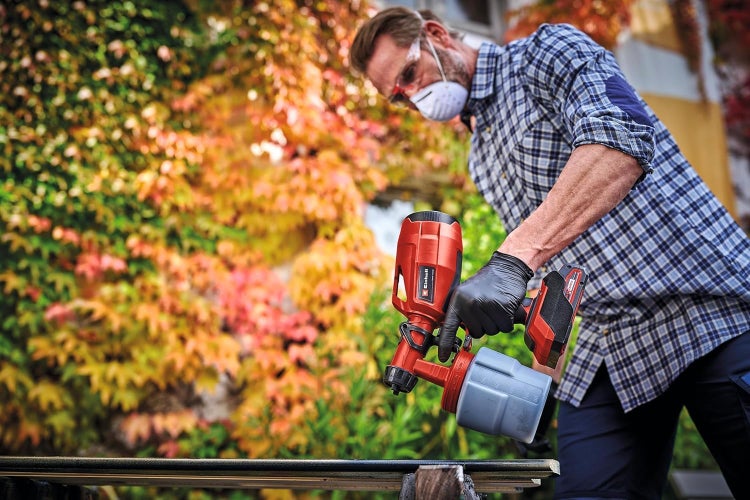 Handwerker verwendet Einhell Akku Farbsprühsystem zum Lackieren eines Holzbretts im Garten vor einer herbstlichen Laubwand mit Schutzausrüstung.