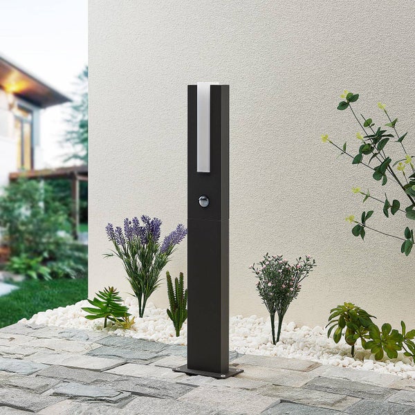 Moderne Außen-Pollerleuchte in Anthrazit mit Bewegungsmelder auf einer Steinterrasse neben einem Gartenbeet mit Kies und Pflanzen.