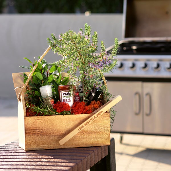 Geschenkkorb mit Kräutern, Soßen und Grillzubehör vor einem Grill