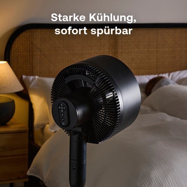 Rückseite eines schwarzen Ventilators in einem Schlafzimmer neben einem Bett mit Geflecht-Kopfteil und weißer Bettwäsche.