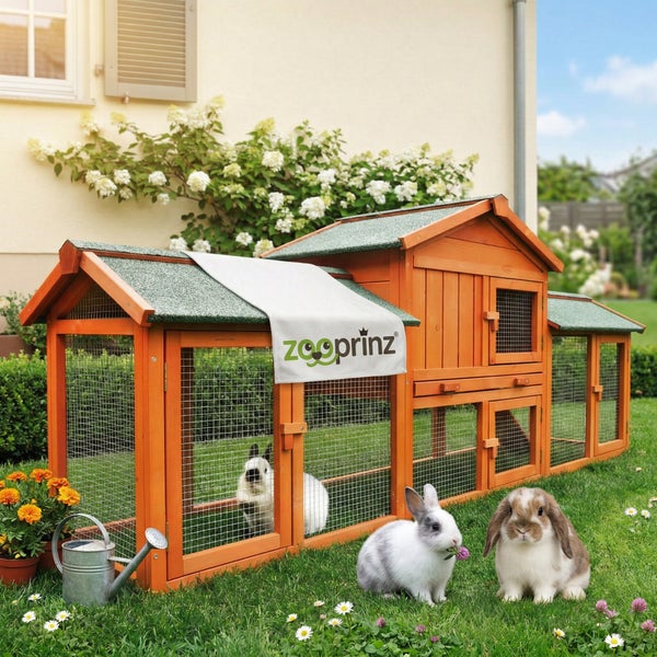 Heller zweistöckiger Kleintierstall aus Holz mit integriertem Auslaufgehege und drei Kaninchen auf einer Rasenfläche. Zooprinz Logo.