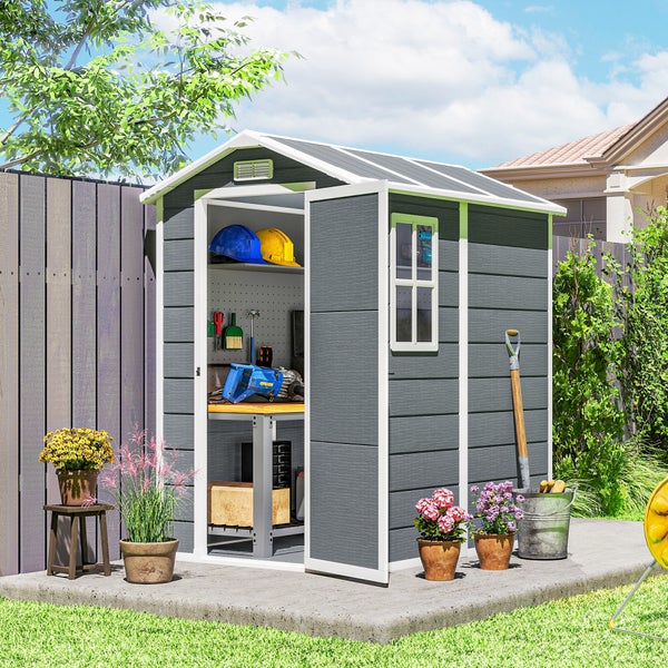 Graues Gerätehaus aus Kunststoff mit weißem Rahmen, geöffneter Tür, Werkbank im Inneren und Blumenkübeln auf einer Betonplatte im Garten.
