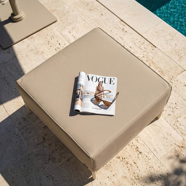 Quadratischer beiger Outdoor-Hocker auf einer Steinterrasse am Pool, dekoriert mit einer Zeitschrift und Sonnenbrille.
