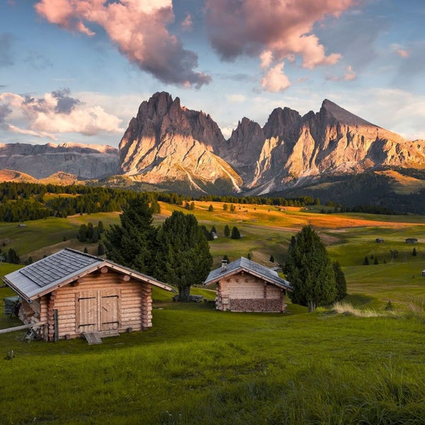 Landschaft mit zwei Holzhütten auf einer Wiese vor einer Bergkette