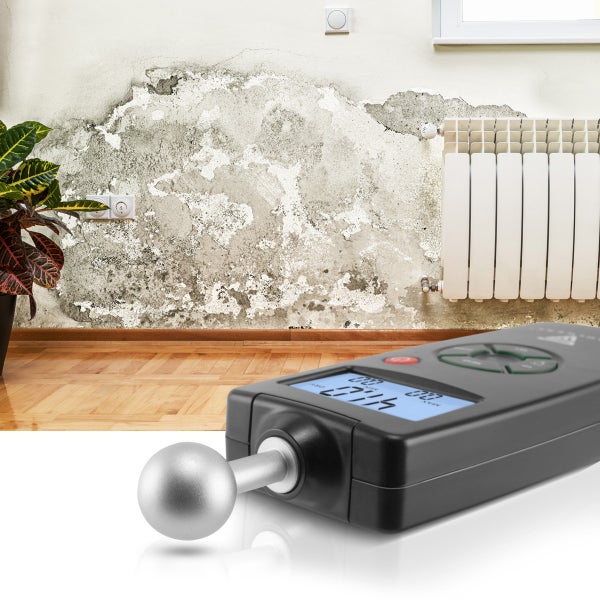 Digitales Feuchtigkeitsmessgerät mit Kugelkopf vor einer feuchten Wand mit abblätternder Farbe und Schimmel neben einem Heizkörper.