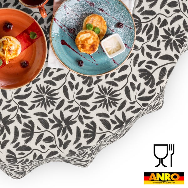 Abwaschbare Tischdecke mit floralem Muster aus Blüten und Blättern, präsentiert mit Frühstücksgeschirr. ANRO DEUTSCHE MANUFAKTUR Logo und lebensmittelecht Symbol.