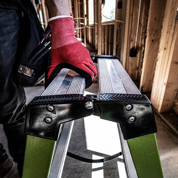 Handwerker mit Arbeitshandschuhen hält eine zusammengeklappte Mehrzweckleiter auf einer Baustelle mit Holzständerwerk.