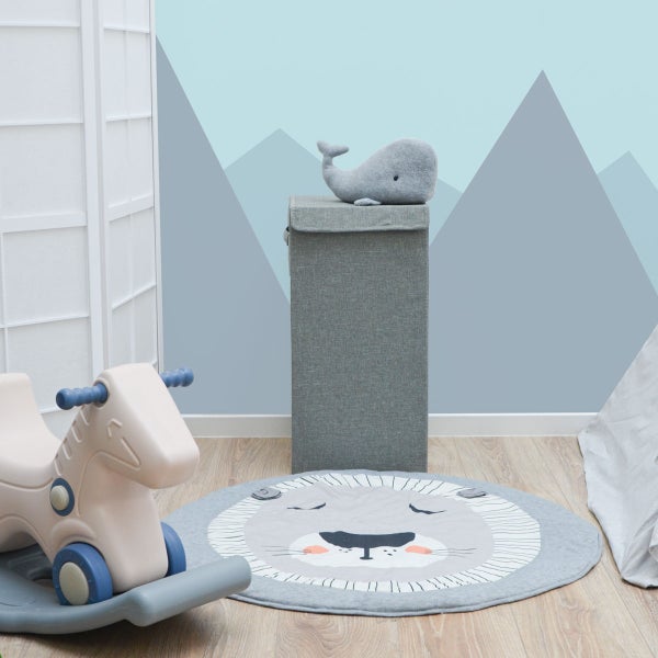 Kinderzimmer mit hoher Aufbewahrungsbox aus Stoff, einem runden Spielteppich mit Löwenmotiv, einem Schaukelpferd und Wandgestaltung mit Bergmotiven.