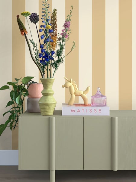 Gestreifte Tapete mit vertikalen Streifen in Beige und Creme hinter einer hellgrünen Kommode mit Blumen und Dekoration.