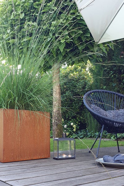Moderner Außenbereich mit einem Pflanzkübel aus Cortenstahl, hohen Ziergräsern, einem schwarzen Loungesessel und einer Laterne auf einer Holzterrasse.