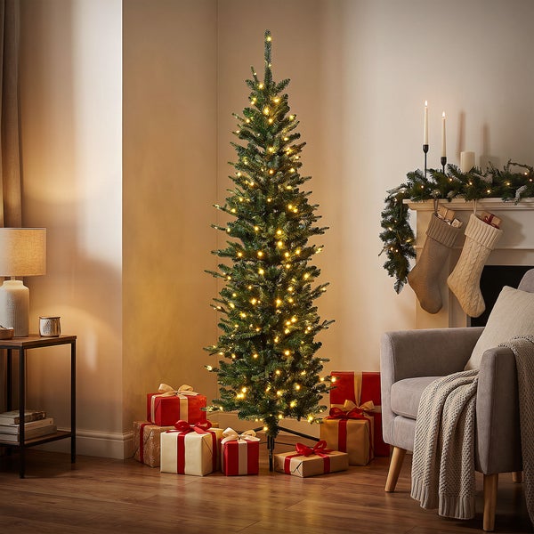 Schlanker künstlicher Weihnachtsbaum mit integrierter LED Beleuchtung, dekoriert mit Geschenkpaketen in einem gemütlich eingerichteten Wohnzimmer.
