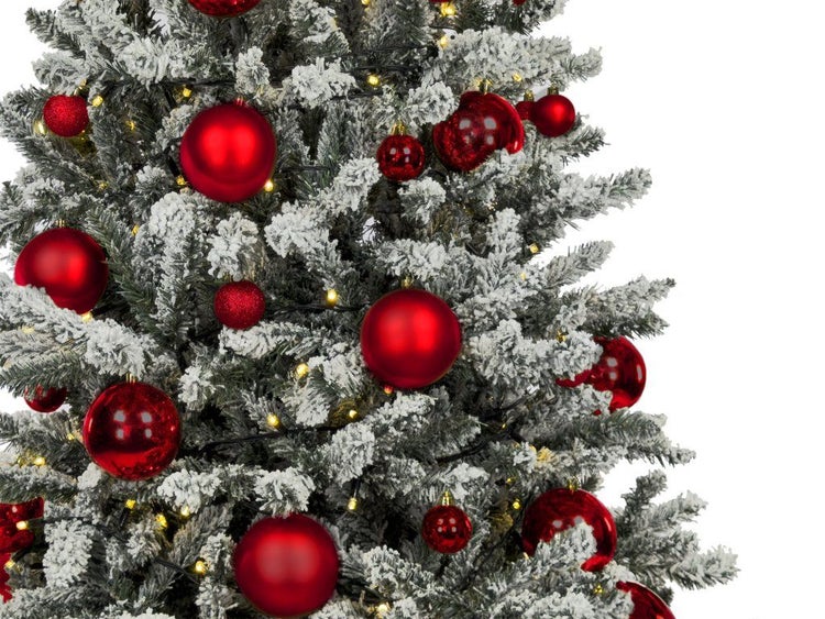 Detailansicht eines verschneiten künstlichen Weihnachtsbaums geschmückt mit roten Christbaumkugeln und einer Lichterkette mit warmweißem Licht.