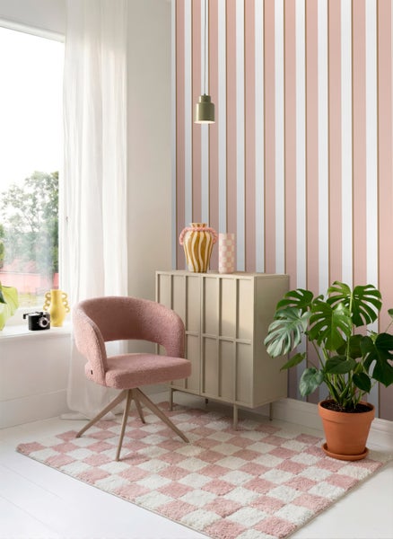 Hell eingerichteter Raum mit gestreifter Vliestapete in Rosa, Weiß und Gold, einem rosa Sessel, kariertem Teppich und einem beigen Sideboard.