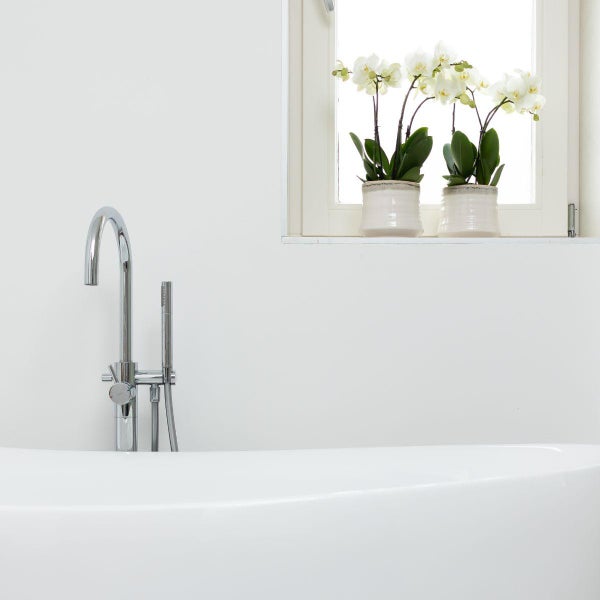 Freistehende Badewannenarmatur aus Chrom mit Handbrause neben einer Badewanne in einem hellen Badezimmer mit Fenster und Orchideen.