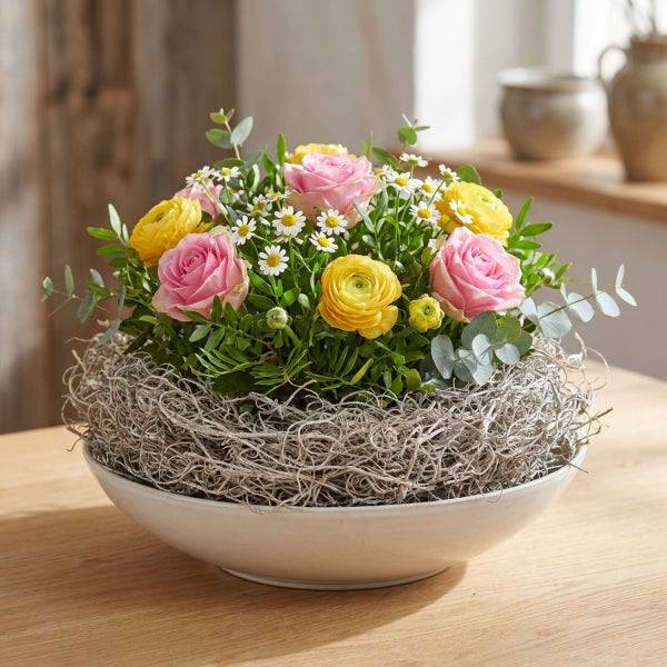 Blumengesteck in einer Schale mit Rosen, Ranunkeln und Margeriten, umgeben von Naturmaterialien auf einem Holztisch.