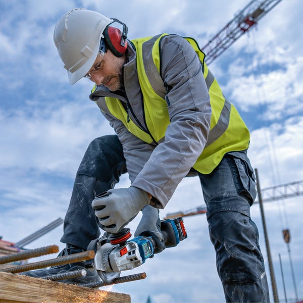 Bauarbeiter nutzt Winkelschleifer auf einer Baustelle, trägt Helm, Gehörschutz und Warnweste beim Schneiden von Metallstäben.