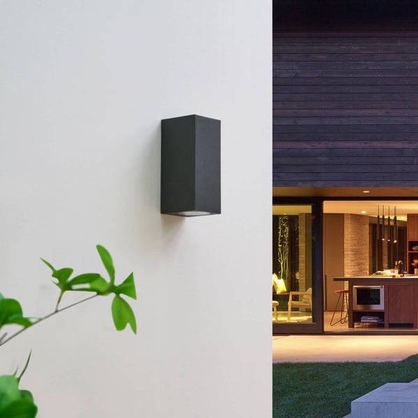 Rechteckige schwarze Außenwandleuchte an einer Fassade neben einer modernen Terrasse mit Blick in eine hell erleuchtete Küche.