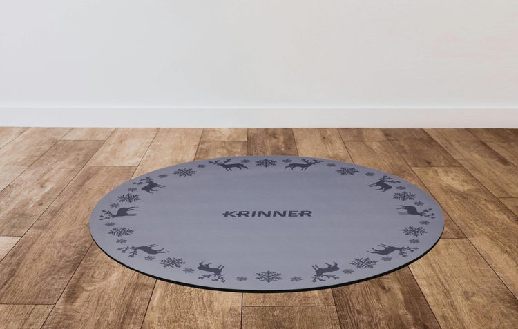 Runde, graue Christbaumunterlage mit Krinner Logo sowie Rentier- und Schneeflockenmotiven auf einem Holzboden.