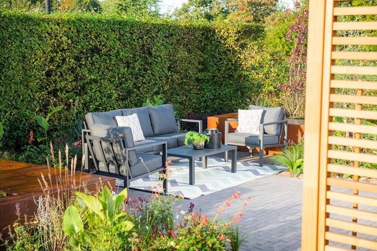 Modernes Garten-Lounge-Set bestehend aus Sofa, Sessel und Couchtisch in Dunkelgrau auf einem Teppich vor einer grünen Hecke.