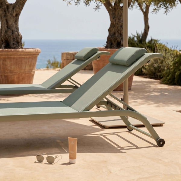 Zwei moderne olivgrüne Sonnenliegen mit Kopfstützen und Rollen auf einer Terrasse vor dem Meer.