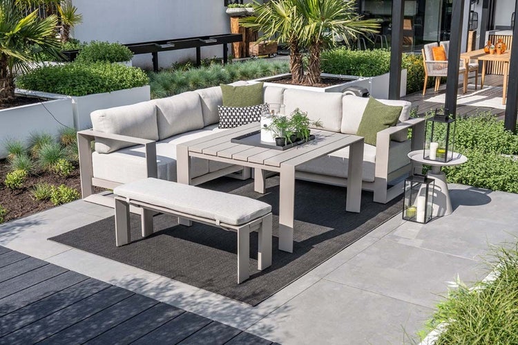 Modernes Garten-Lounge-Set aus Metall mit Ecksofa, Esstisch, Sitzbank und Outdoor-Teppich auf einer Terrasse mit Bepflanzung.