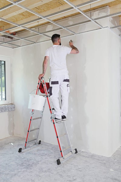 Ein Mann steht auf einer Leiter und verputzt eine Wand in einem Raum mit abgehängter Decke.