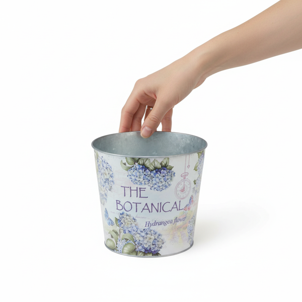 Hand greift in einen Metallübertopf mit Hortensien-Druck und dem Schriftzug The Botanical Hydrangea flower.