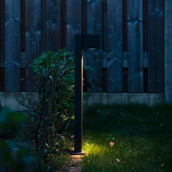 Moderne Wegeleuchte aus Metall beleuchtet eine Rasenfläche und einen Busch in einem Garten vor einem Holzzaun bei Nacht.