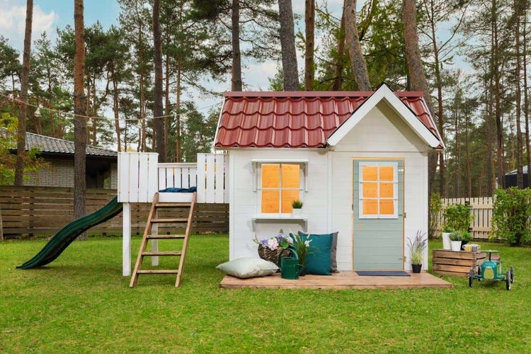 Weißes Spielhaus aus Holz mit rotem Ziegeldach, grüner Rutsche, Leiter und kleiner Veranda in einem Garten mit Bäumen.