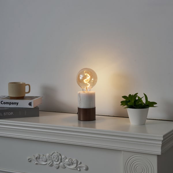 Moderne Tischleuchte mit Marmorsockel und eingeschaltetem Dekoleuchtmittel auf einer weißen Kommode mit Büchern und einer Pflanze.