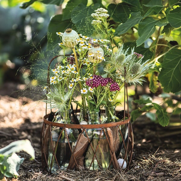 Blumenarrangement in Glasvasen in einem runden Metallkorb, stehend im Garten, dekoriert mit Mohnkapseln, Schafgarbe und verschiedenen Wiesenblumen.