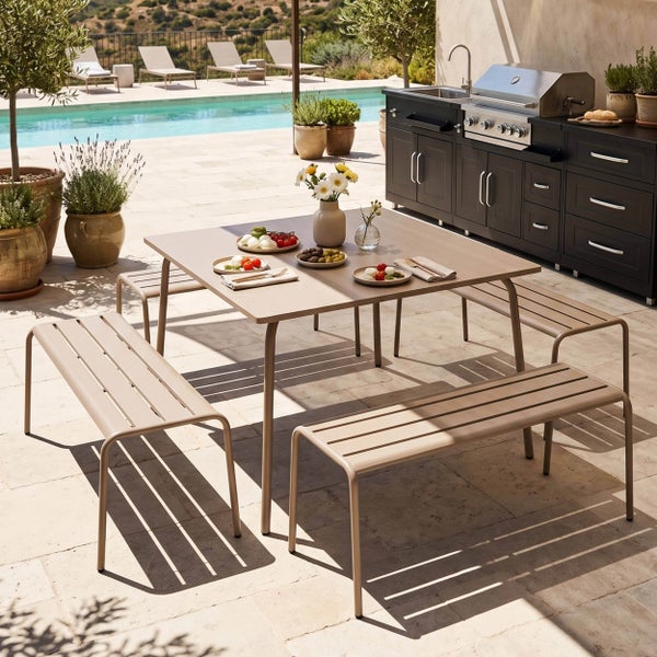 Gartentisch mit Sitzbänken aus Metall auf einer Terrasse neben einer Außenküche mit Grill und einem Pool im Hintergrund. Hell und modern.