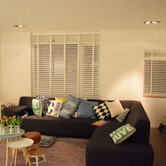 Modernes Wohnzimmer mit großem dunklen Ecksofa, verschiedenen Kissen und weißen Jalousien an den Fenstern.