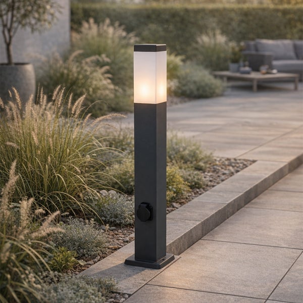 Moderne Außenleuchte in Anthrazit mit integrierter Steckdose auf einer Terrasse neben Ziergräsern im Garten.