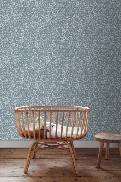 Kinderzimmer mit einem Babybett aus Rattan und einem Holzhocker vor einer blauen Tapete mit weißem Blumenmuster und dunklem Holzboden.
