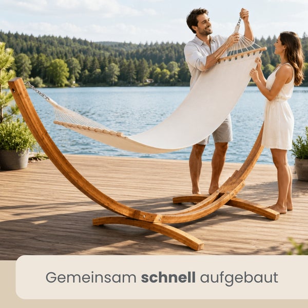 Ein Paar montiert eine helle Hängematte an einem bogenförmigen Holzgestell auf einer sonnigen Holzterrasse an einem See.