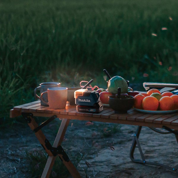 Szene mit vollem Holztisch im Freien, darauf Geschirr, Obst und eine Makita Akku-Lampe