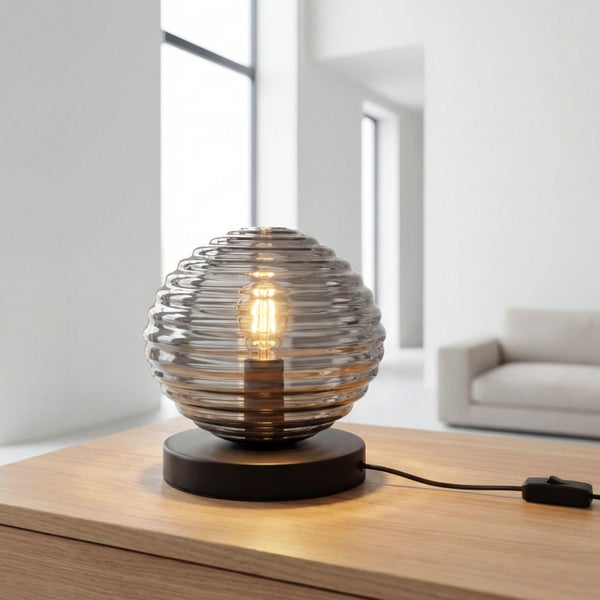 Moderne Tischleuchte mit geriffeltem, rauchfarbenem Glasschirm auf einem Holztisch in einem hellen, aufgeräumten Wohnzimmer.