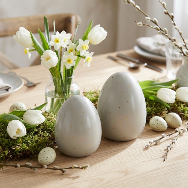 Osterdekoration mit zwei eiförmigen Dekofiguren mit goldenen Punkten auf einem Holztisch mit Tulpen, Narzissen und Weidenkätzchen.