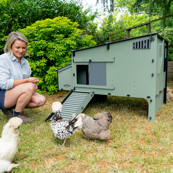 Heller Garten mit erhöhtem Hühnerstall aus Recycling-Kunststoff. Frau hockt neben Hühnern vor dem Stall mit Rampe und Schiebetür.