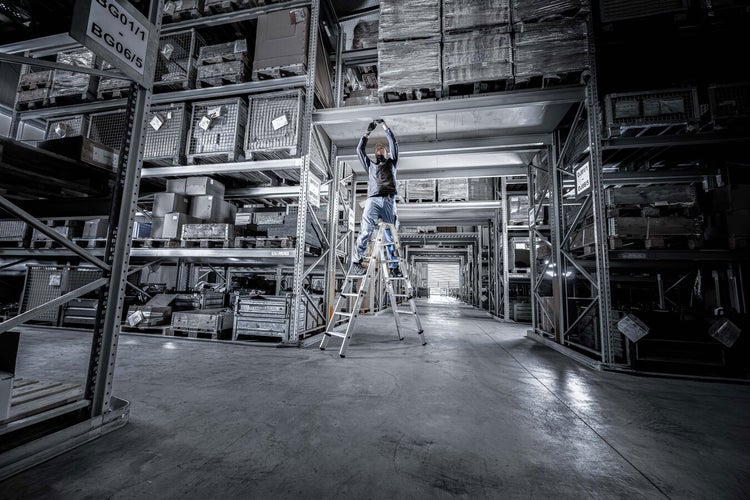 Eine Person steht auf einer Trittleiter in einem Lagerhaus mit Regalen voller Kisten und Waren.