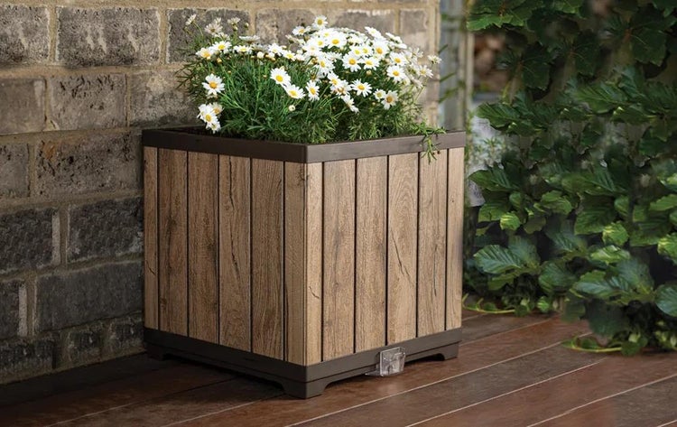 Quadratischer Pflanzkübel in Holzoptik mit weißen Margeriten auf einer Holzterrasse vor einer Steinmauer.