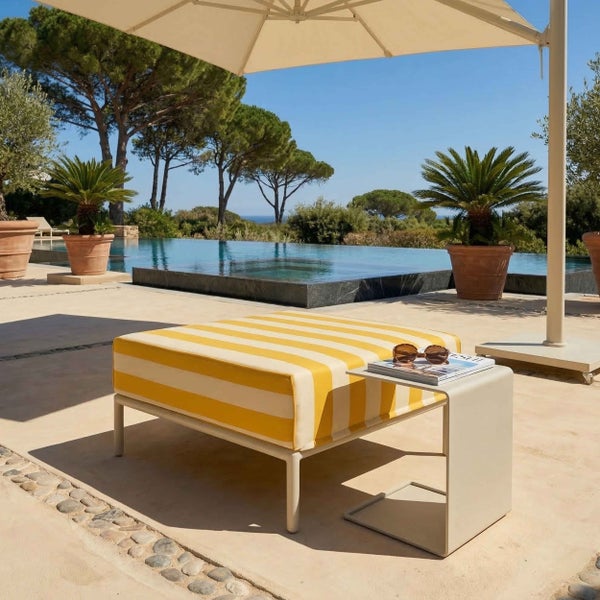 Gestreifter Hocker und Beistelltisch auf einer hellen Poolterrasse mit Palmen und Sonnenschirm.