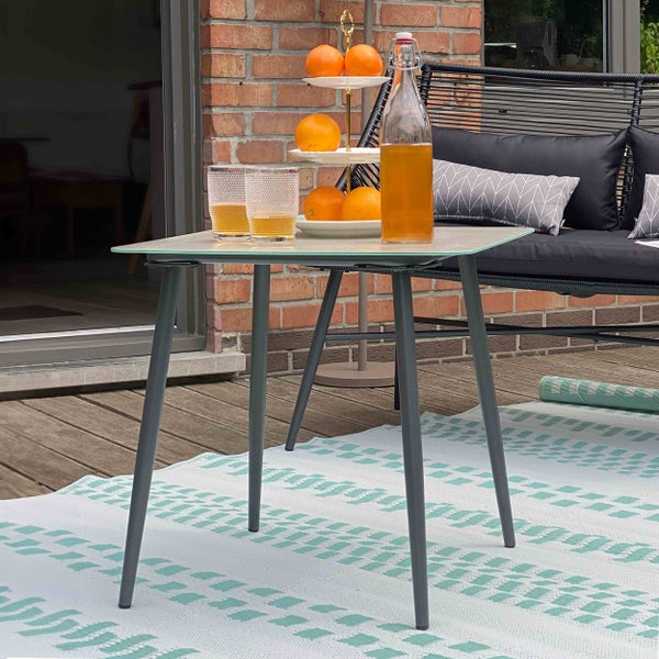 Quadratischer Gartentisch mit dunklen Metallbeinen und heller Platte auf einer Terrasse mit Outdoorteppich und Lounge Sofa.