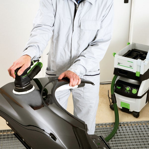 Ein Handwerker schleift ein schwarzes Autoteil mit einem Exzenterschleifer von Festool, während ein Staubsauger daneben steht