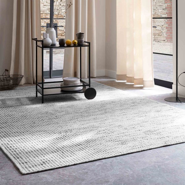 Hellgrauer gewebter Teppich in einem hellen Wohnzimmer mit schwarzem Servierwagen und hellen Vorhängen.