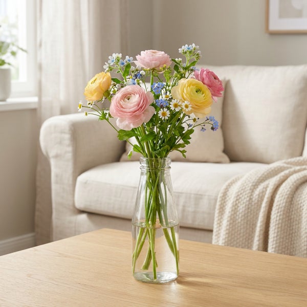 Blumenstrauß aus Ranunkeln und kleinen Frühlingsblumen in einer gläsernen Flaschenvase auf einem hölzernen Couchtisch in einem hellen Wohnzimmer.
