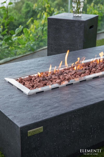 Rechteckiger Gaskamin-Tisch in Steinoptik mit Lavasteinen und Flammen auf einer Terrasse.