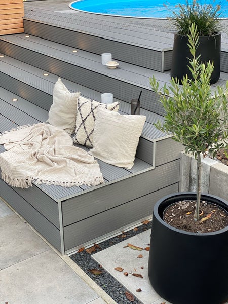 Hell gestaltete Außentreppe aus Terrassendielen mit Sitzbereich aus Kissen und Decke neben einem Pool und Pflanzkübeln.